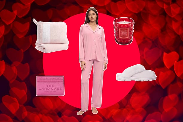 Woman stands against a red heart-patterned background, surrounded by thoughtful Valentine's Day gifts for her: a pink travel blanket, red candle, pink card case, cozy pink slippers and pink pajamas that she's wearing