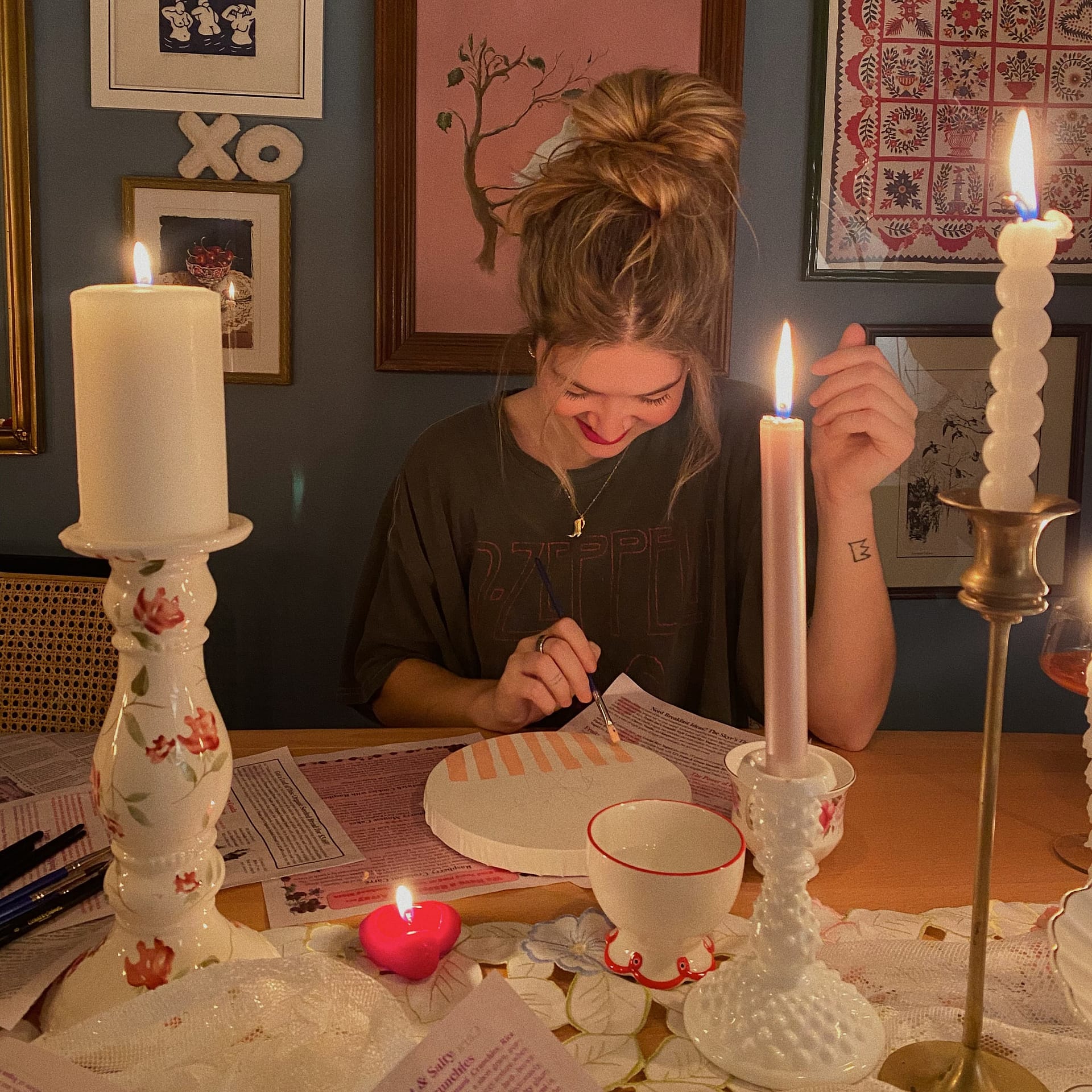 Woman with a bun hairstyle paints a circular object at a candle-lit table surrounded by framed artwork on the wall
