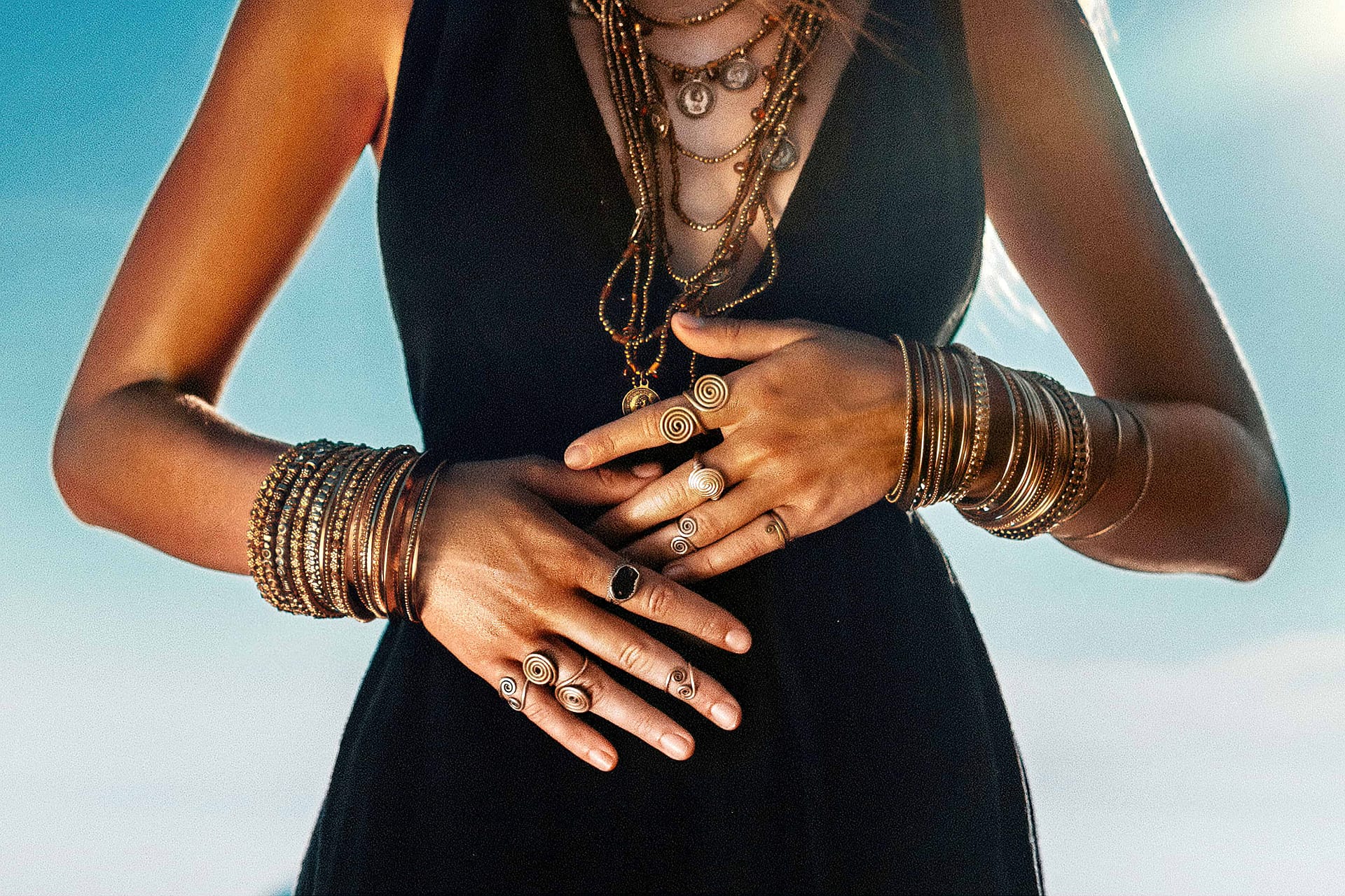 Woman in a black dress showcasing numerous bracelets, rings, and necklaces, with hands adorned in shimmering bracelets placed above the waist against a blue sky background